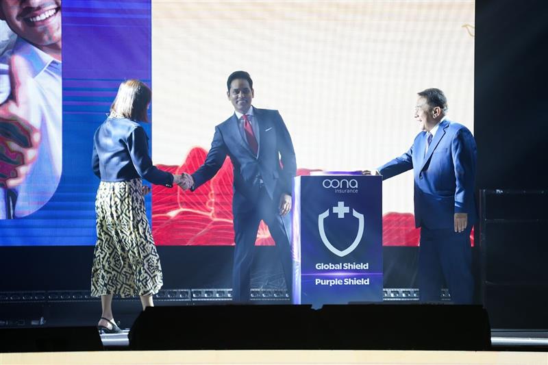Two executives shaking hands on stage beside an Oona Insurance podium displaying Global Shield and Purple Shield during a healthcare protection presentation.