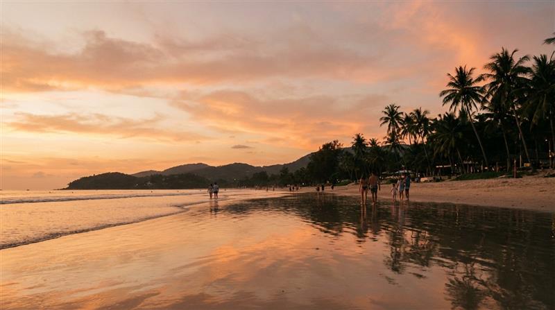 Families and couples strolling along Kata Beach Phuket Thailand at sunset.